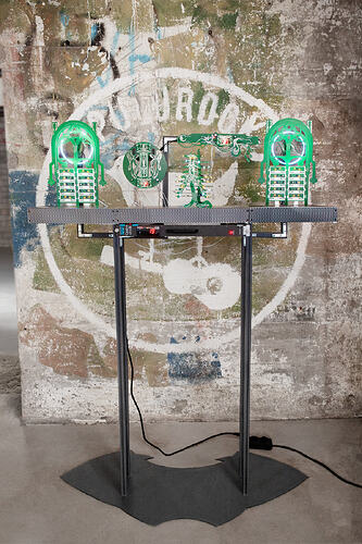 Slack Storage, circuit boards on a server room cooling fan tray - Front view of the full installation on a black stand. Two symmetrical green board figures with glowing white rings and stacked lit modules flank a circular board and a rib-cage form, with an arched board across the top. A rack unit on the table shows a red '19' display. A faded circular painting marks the concrete wall behind.