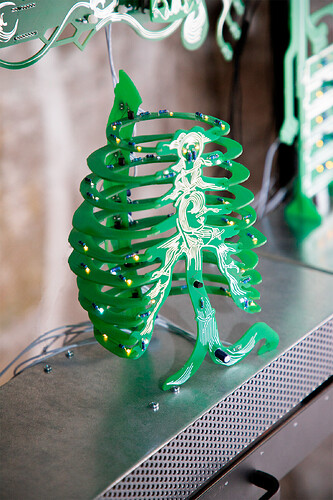 Slack Storage, rib cage. A green circuit-board sculpture resembling a rib cage and spine: curved horizontal ribs stacked around a central figure in white line-work. Small yellow and blue LEDs light the ribs. It stands on a perforated metal surface with more board structures behind.
