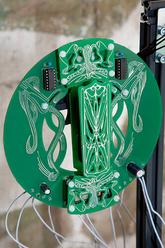 Slack Storage, ribboned coil. A circular green circuit board covered in symmetrical white line-work, with two black DIP chips near the top, electrolytic capacitors and white cabling. It is mounted on a black aluminium-profile frame against a concrete wall.